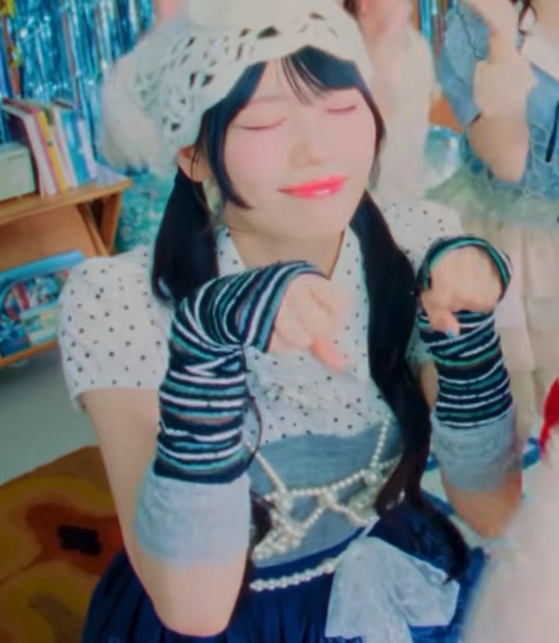 A young woman in a blue polka-dot outfit with striped arm warmers and a lace headpiece smiles with her eyes closed in a pastel-themed room.