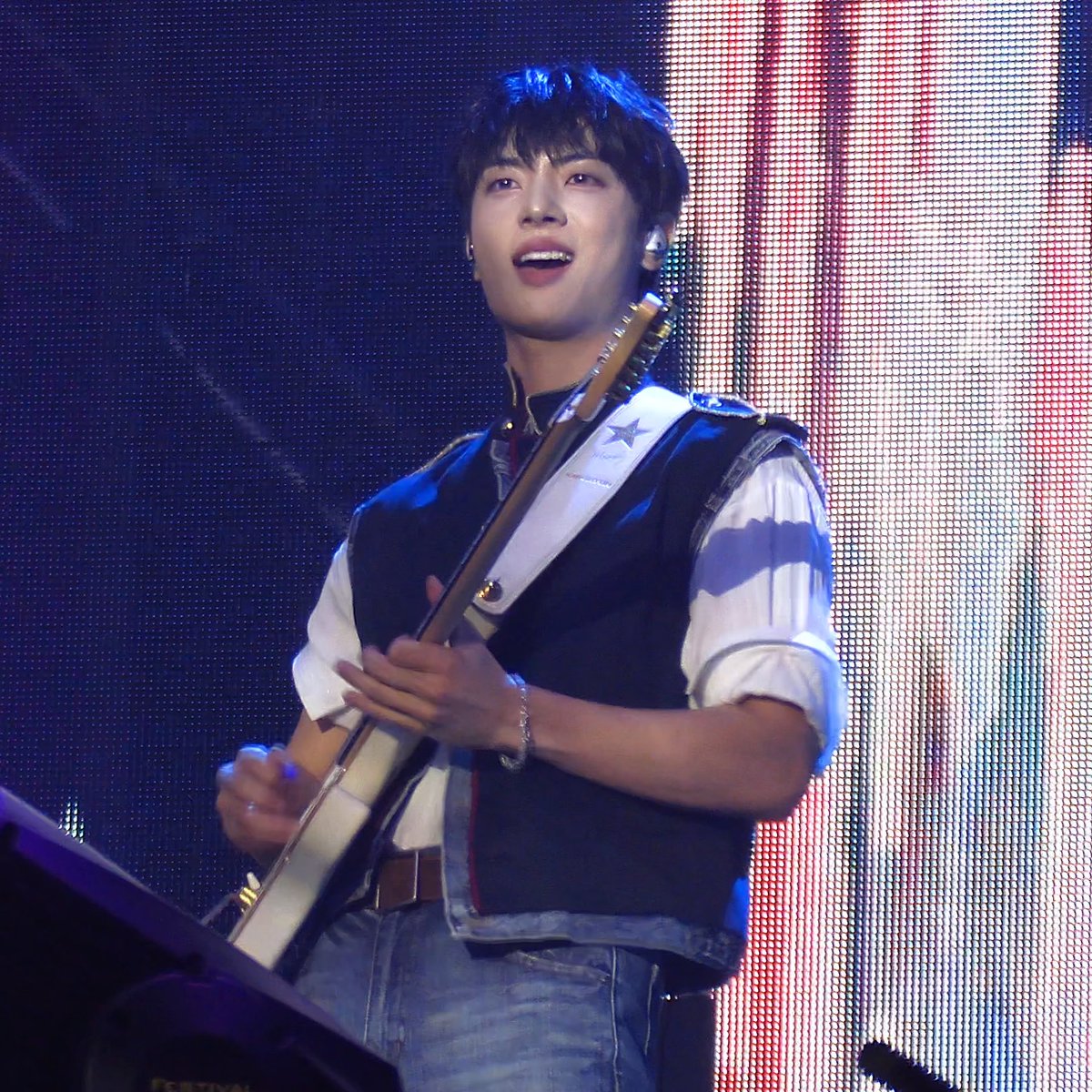A young male musician performs on stage, playing a guitar and singing while wearing a white shirt with a dark vest.