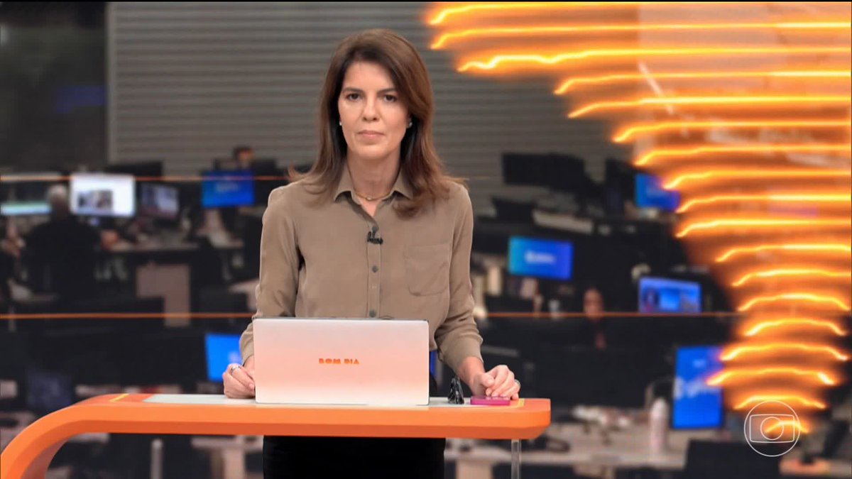 A female news anchor with brown hair wearing a beige shirt stands at a desk with an orange top, holding a white laptop with "SAMSUNG" written on it in a newsroom setting.