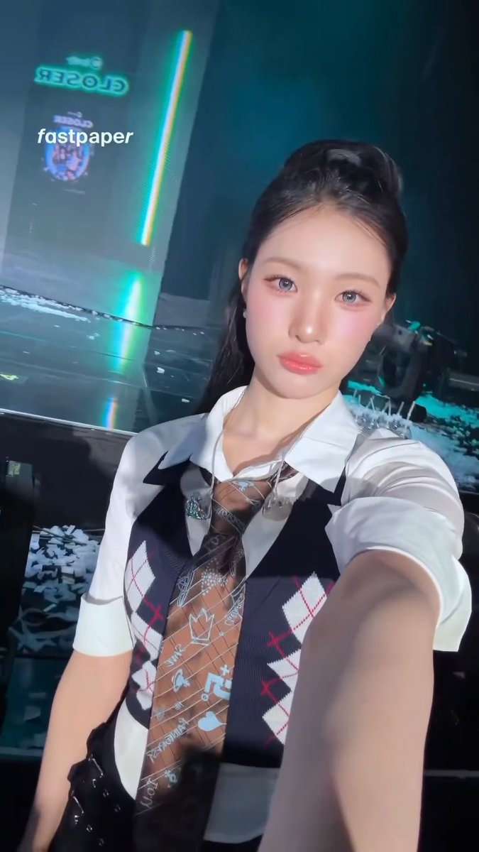 A selfie of a young woman with long dark hair wearing a white collared shirt with an argyle vest and tie, taken at what appears to be a concert venue with a neon sign in the distance; the text 'fastpaper' is visible.