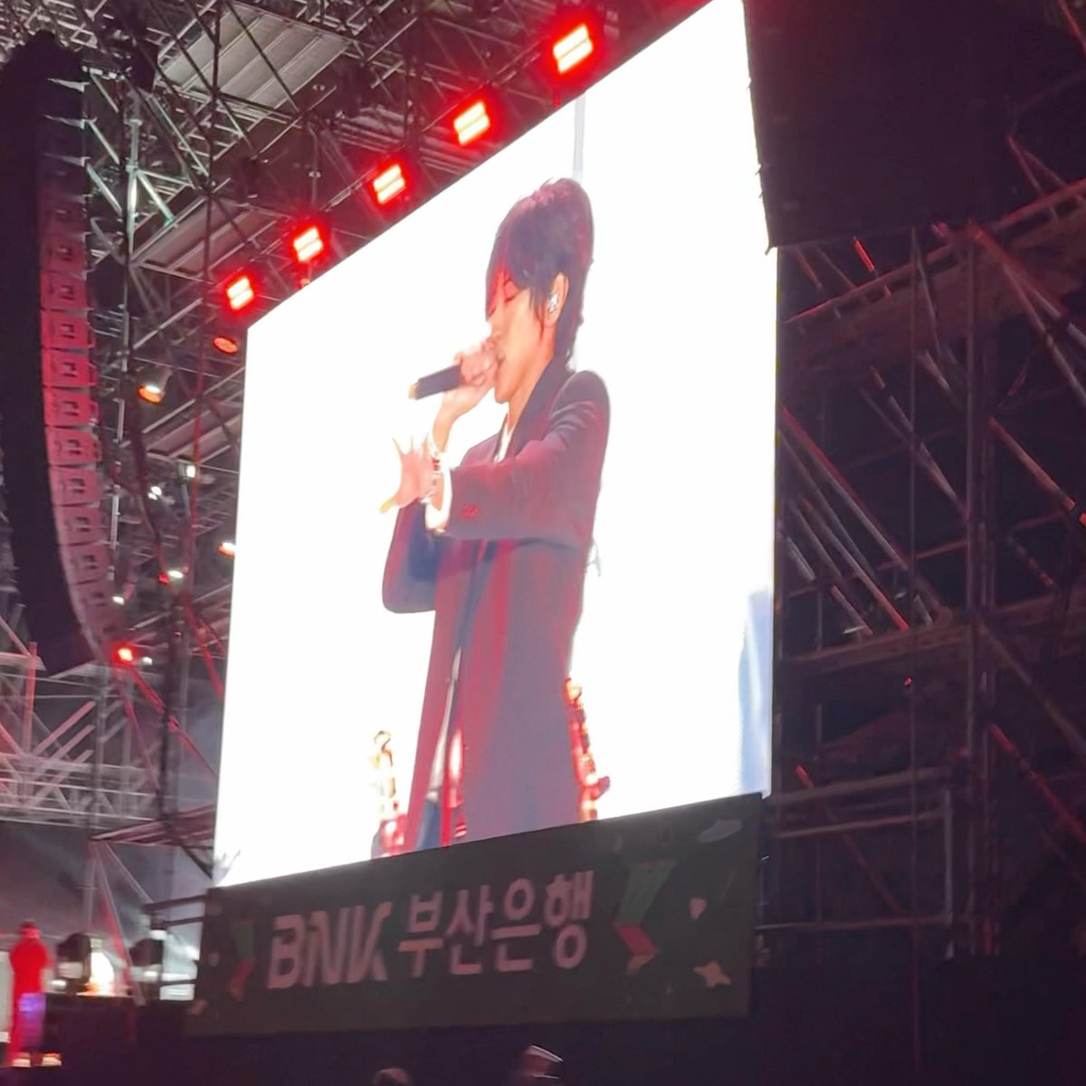 A performer, identified as WINNER's Kang Seungyoon, sings into a microphone while being displayed on a large concert screen, with a banner below reading "BNK 부산은행" (BNK Busan Bank) suggesting the event is sponsored by the bank. The image captures the energetic live performance referenced in the post, where Seungyoon's on-stage antics and crowd chants of "dance dance dance dance" provided an unforgettable hangover cure.