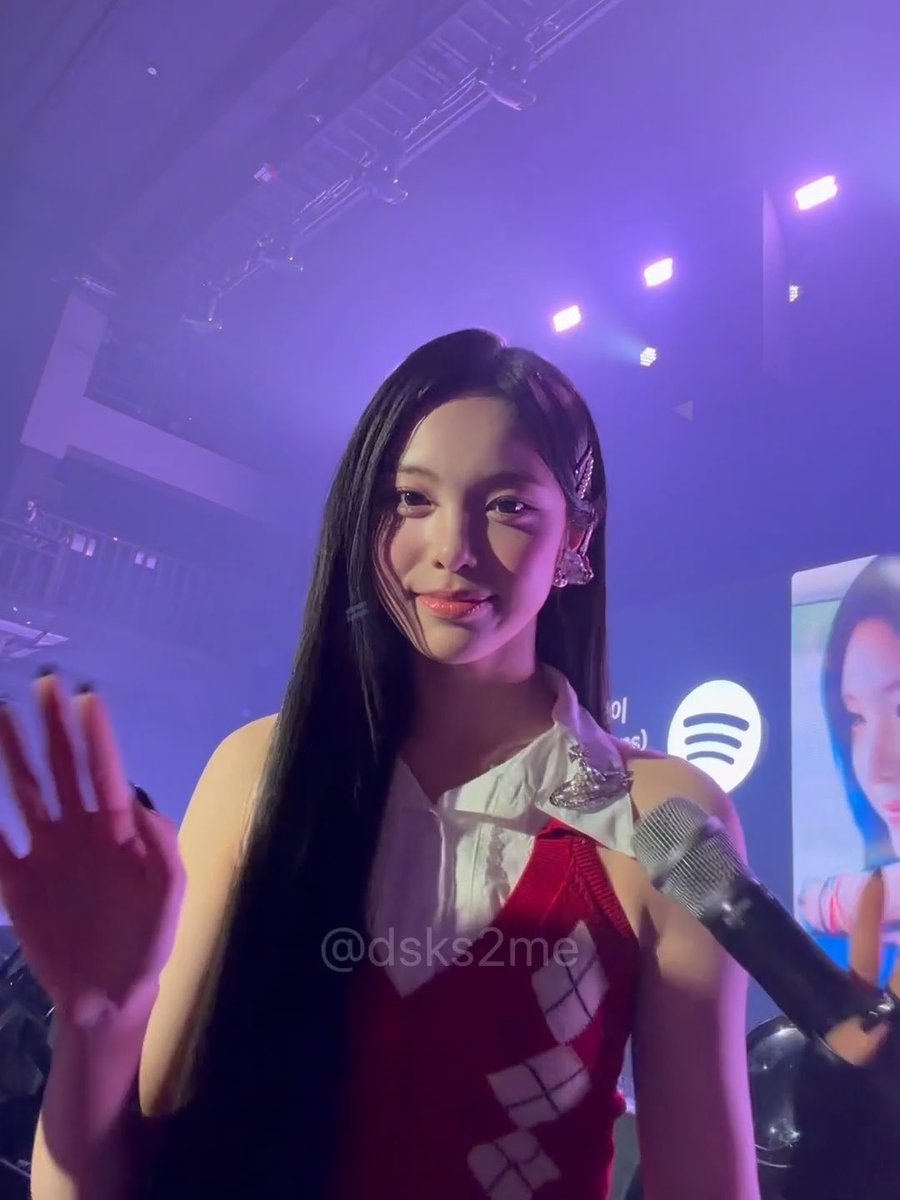 A performer with long dark hair and a red argyle dress waves at the camera while holding a microphone on a stage with purple lighting and a Spotify backdrop. The image, tagged "#이안" (Lee An), captures the singer looking serene and angelic as she greets fans at what appears to be a live event.