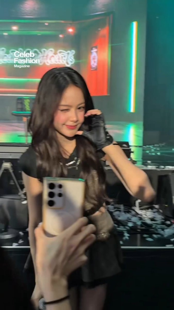 A young woman with long dark hair smiles while taking a selfie in what appears to be a backstage area with neon green and red lighting, wearing a black outfit with fingerless gloves; 'Celeb Fashion Magazine' and 'Neon Bunny' signage is visible in the background.