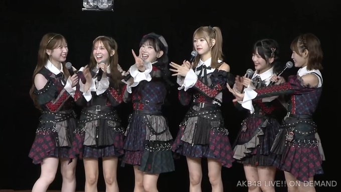 Seven young women in matching black, red, and white outfits performing on stage with microphones, captioned 'AKB48 LIVE!! ON DEMAND'.