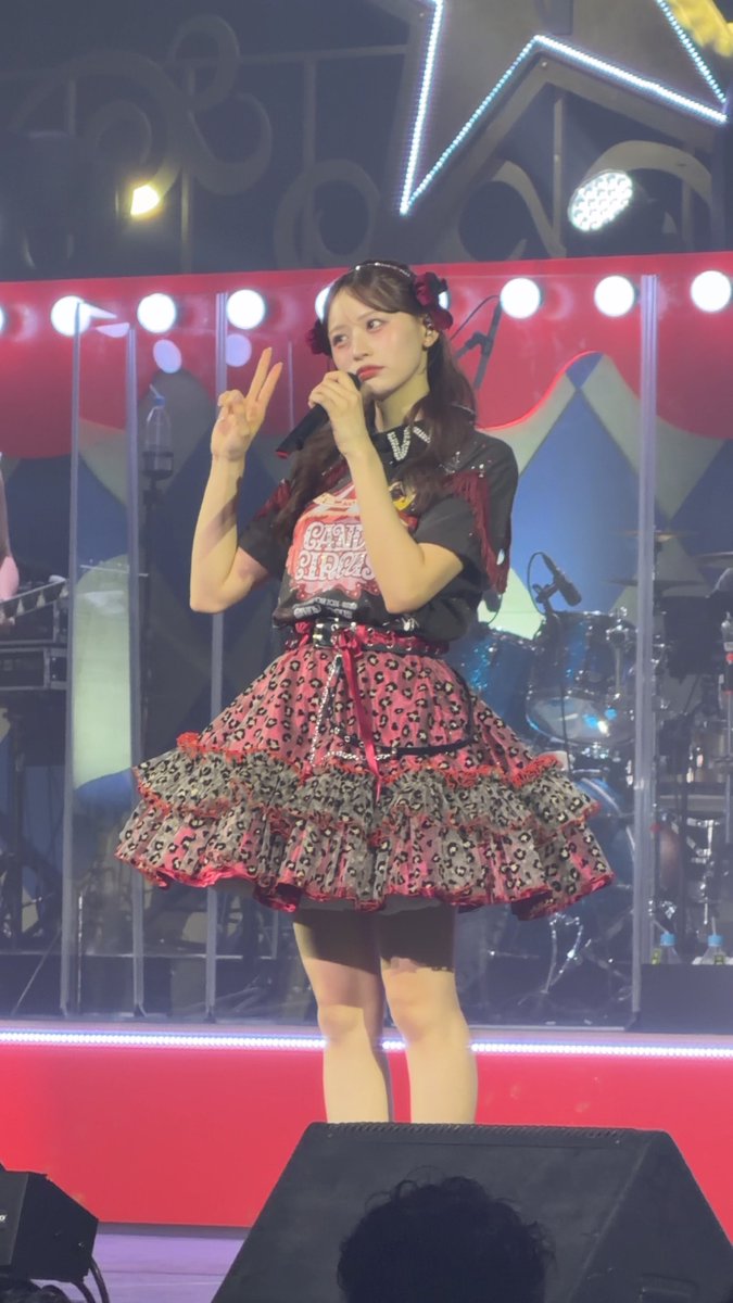 A young woman in a red and black patterned idol dress performing on stage, holding a microphone and making a peace sign.