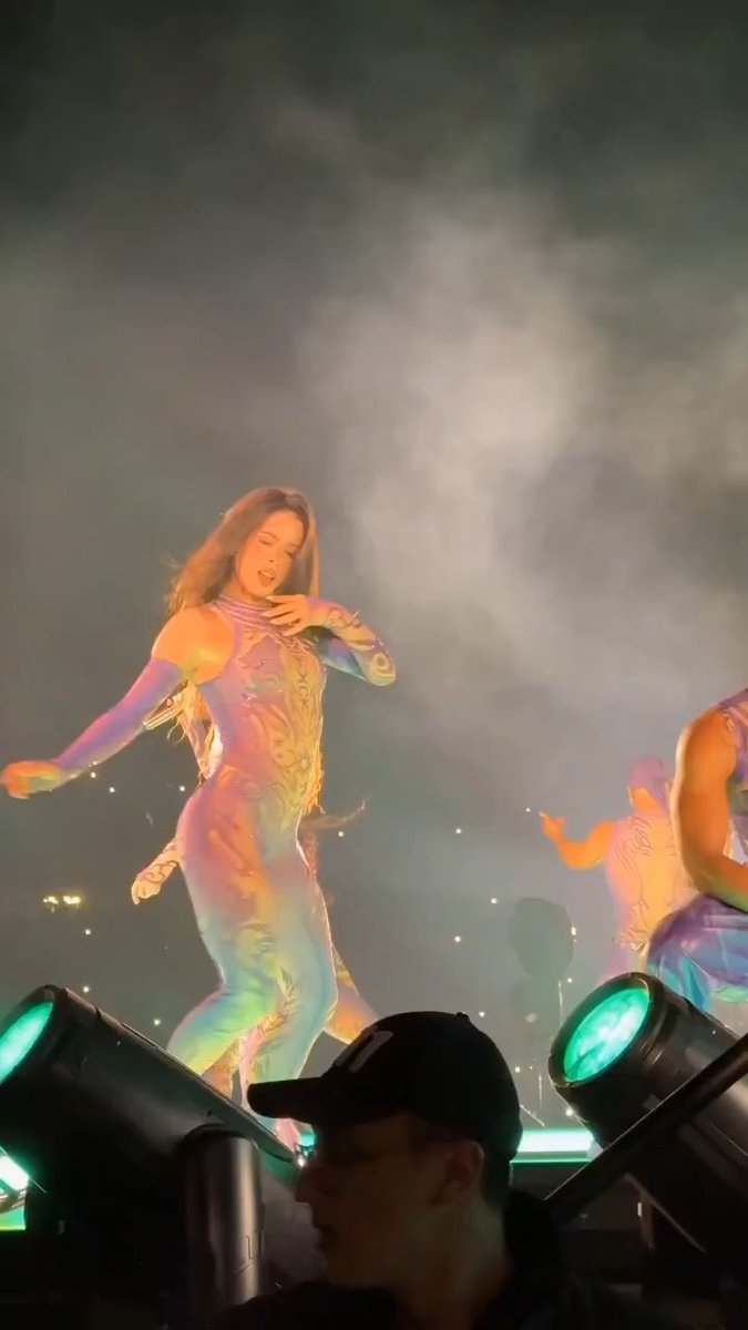A dancer in a holographic, iridescent bodysuit performs on stage under dramatic lighting and smoke, with backup dancers visible behind her. The image relates to a social media comment joking that Shakira would never want to share the stage with such a captivating performer.