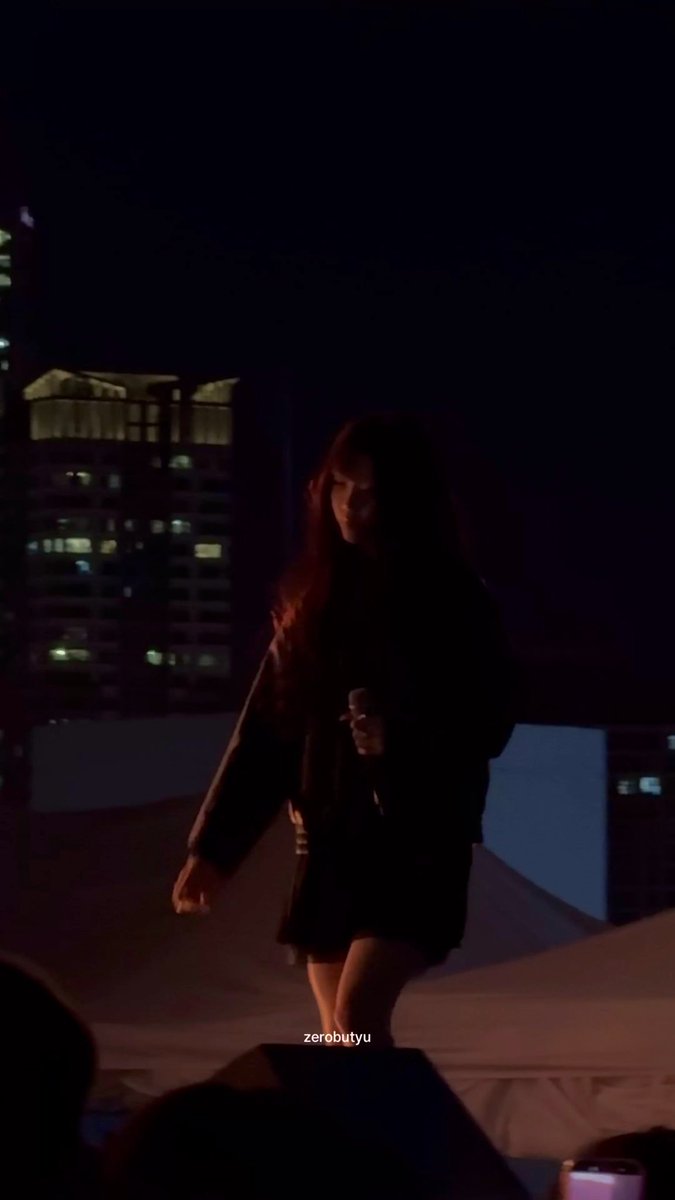 A young woman with long hair stands on a stage at night against a dark cityscape with illuminated high-rise buildings, holding a microphone, with the watermark 'zerobutyu' visible.