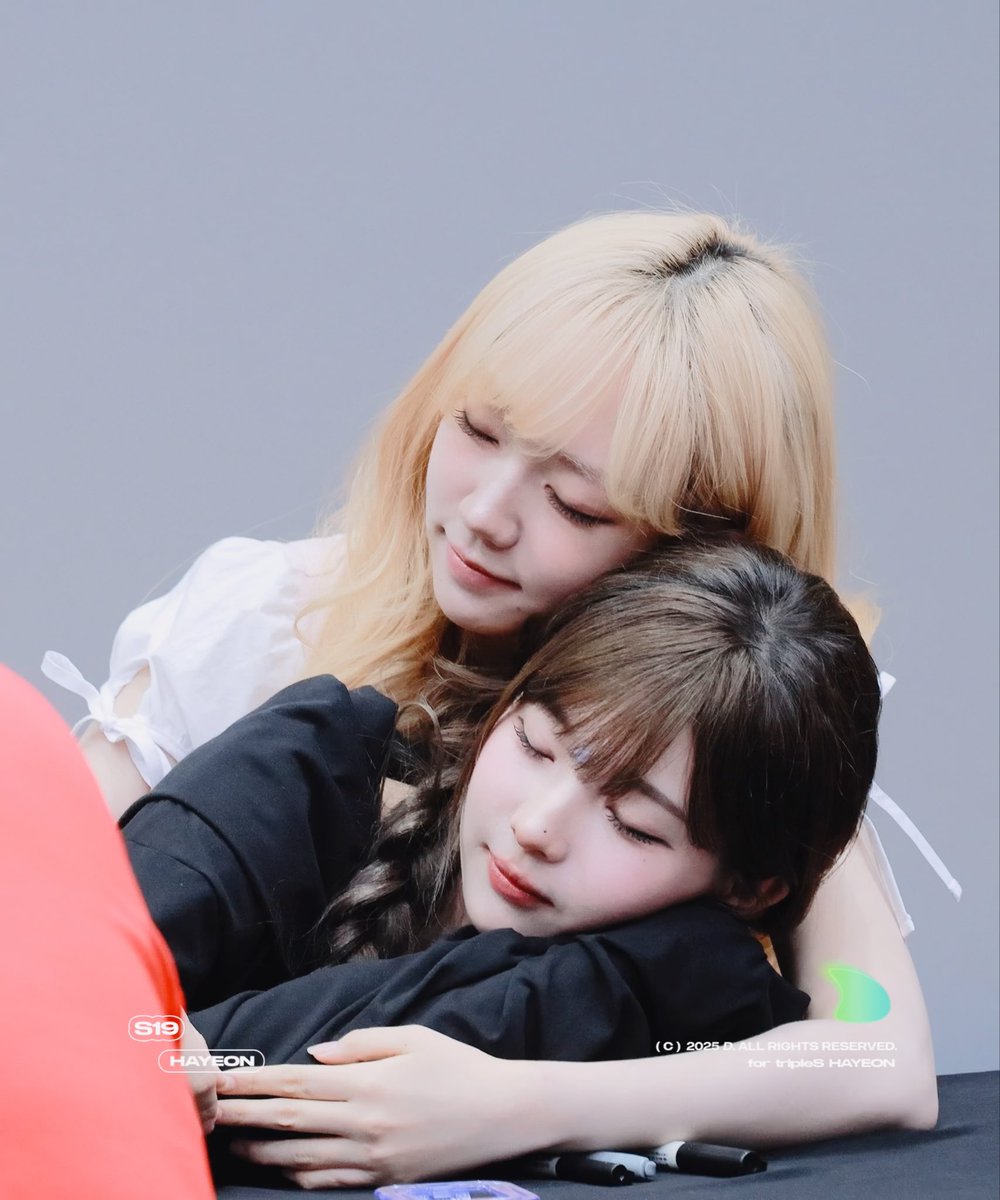 Two young women share an affectionate hug; the blonde woman in white gently rests her chin on the brown-haired woman in black, with watermarks indicating 'S19 HAYEON' and 'tripleS HAYEON'.