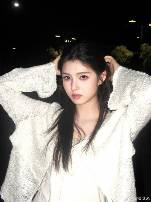 A nighttime portrait of a young woman wearing a white knit sweater, with her hands raised to her hair, looking at the camera with red lipstick.