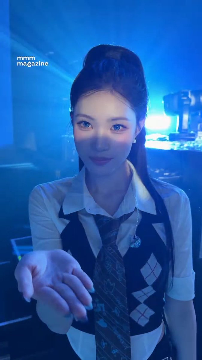 A young woman wearing a white shirt, argyle vest, and dark tie reaches her hand toward the camera in a blue-lit setting, with 'mmm magazine' written in the corner.