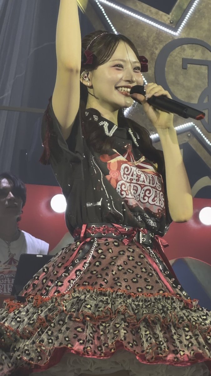A young woman performs on stage holding a microphone and raising one arm, wearing an elaborate dark costume with red accents, a leopard print skirt, and a shirt reading 'CIRCUS CIRCUS'.