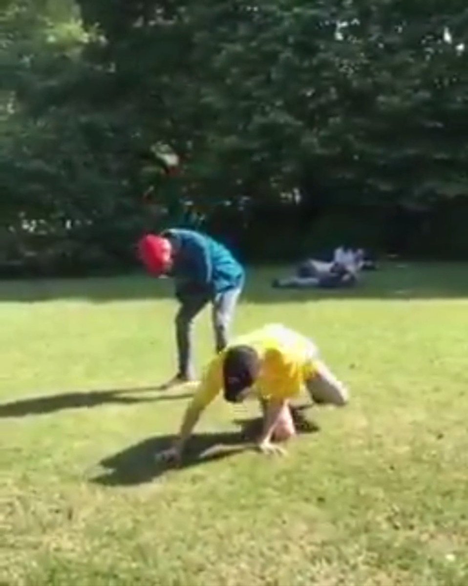 A blurry outdoor scene on a grassy field where a person in a yellow shirt is on all fours while another person in a blue jacket and red cap bends over behind them, with trees and what appears to be a large rock or object in the background.