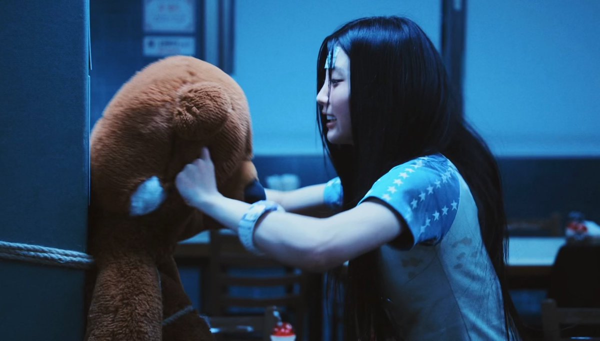 A long-haired young woman wearing a white and blue star-patterned shirt smiles as she touches the snout of a big brown teddy bear in a dimly lit, blue-toned room.