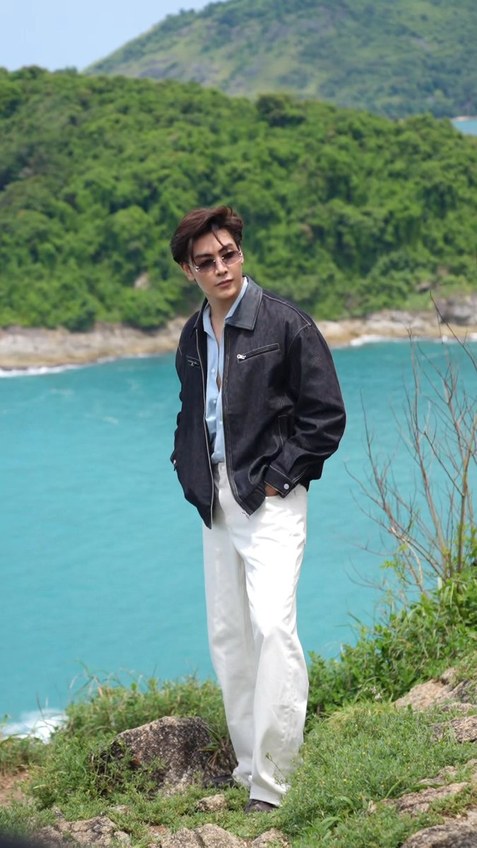 Joong Archen stands on a grassy cliff overlooking a turquoise bay and lush green mountains, wearing a black leather jacket, light blue shirt, white pants, and sunglasses, as part of the "Dr. Ally Together in Paradise with Joong" fan activity event held on September 19-20, 2026.