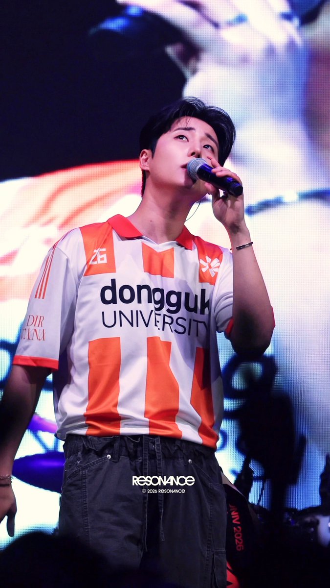 A male performer with dark hair sings into a microphone on stage, wearing an orange and white "dongguk UNIVERSITY" jersey paired with dark "RESONANCE" cargo pants. The image corresponds to the post about 영케이 (Young K) of DAY6 performing at Dongguk University on September 30th, with the caption praising his rhythmic footwork as truly masculine.