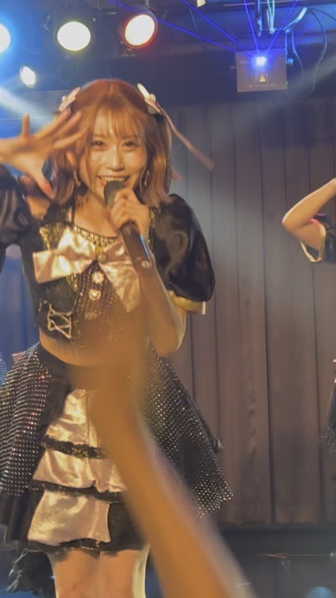 A smiling young woman with orange-brown hair performs on stage under bright spotlights, holding a microphone in one hand while wearing an elaborate black and gold idol-style outfit with a large bow.