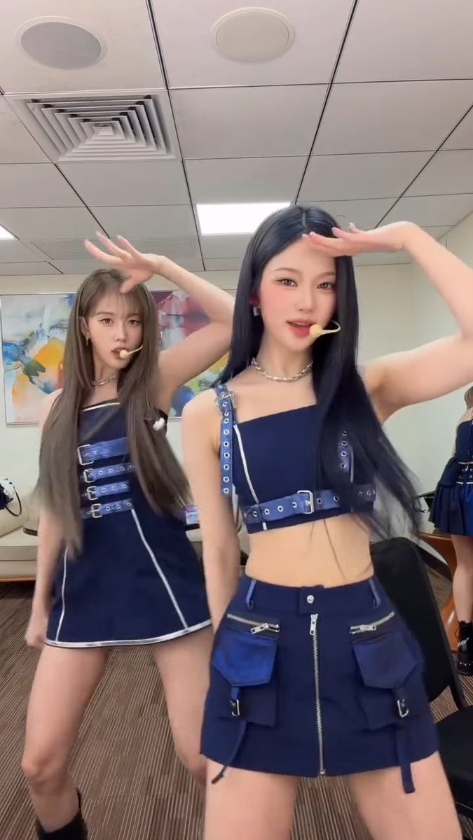 Two female performers in matching navy blue two-piece outfits with buckles and zippers dance in a studio setting, with the woman in front saluting with her hand.