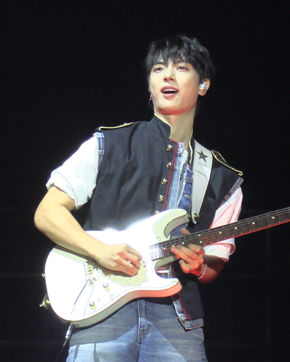 A young man with dark hair performs on stage playing a white electric guitar, wearing a black vest with gold trim over a white shirt and jeans, with an in-ear monitor visible.