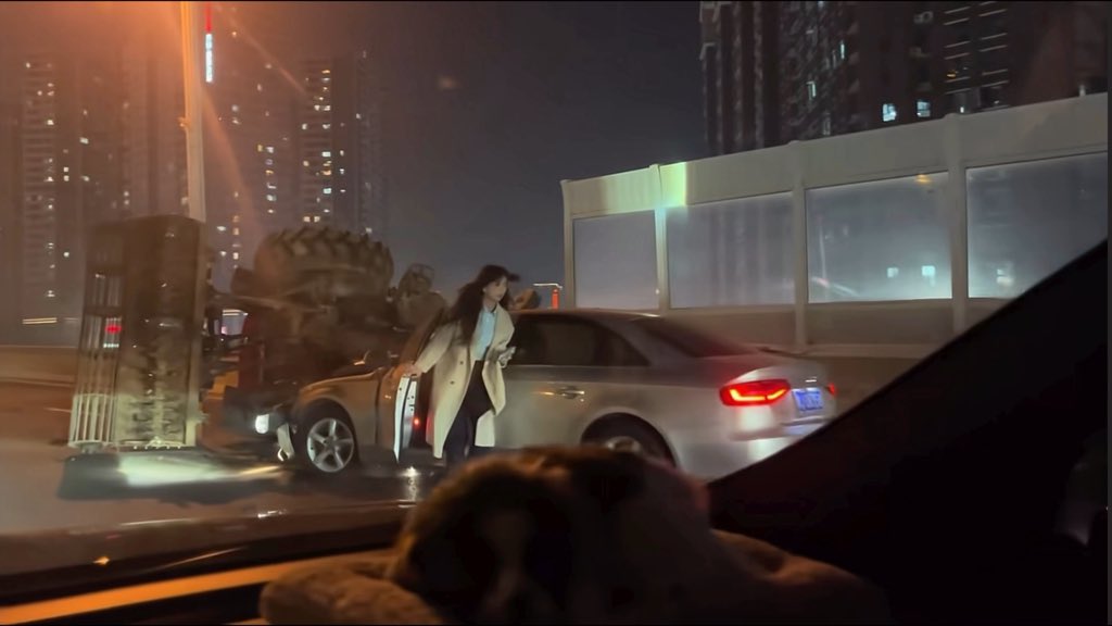 A nighttime street scene showing a woman in a light coat stepping out of a silver Audi sedan parked next to a city overpass, viewed from inside another vehicle.