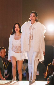 Two women walking together on a runway during a show, dressed in white outfits, one wearing a long white jacket and the other in a white dress.