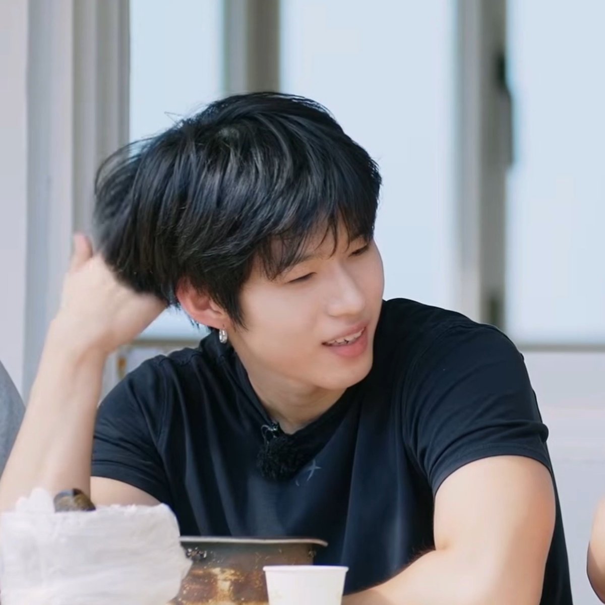 A young man with black hair wearing a black t-shirt smiles and touches his head while sitting at a table.