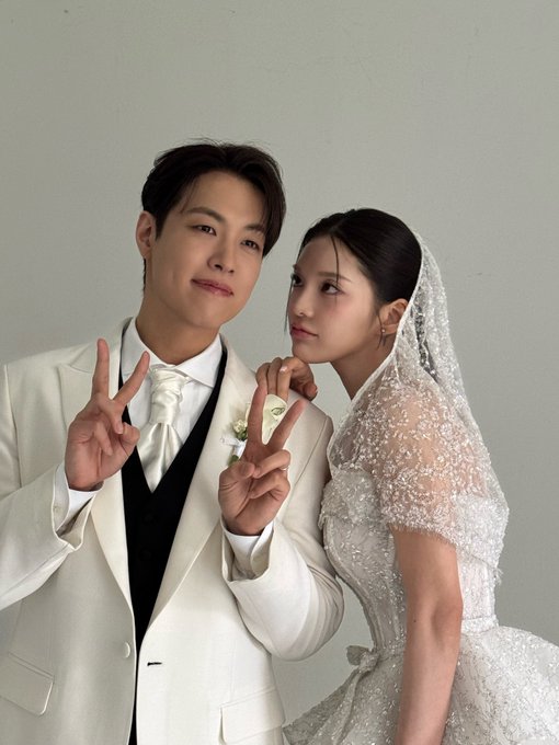 A bride and groom pose together in formal wedding attire; the groom wears a cream suit and the bride wears a beaded lace gown with a veil, both making peace signs.