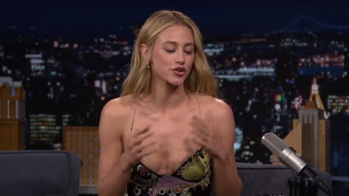 A blonde woman in a floral dress speaks on a late-night talk show set, gesturing with her hands against a backdrop of a city skyline at night.