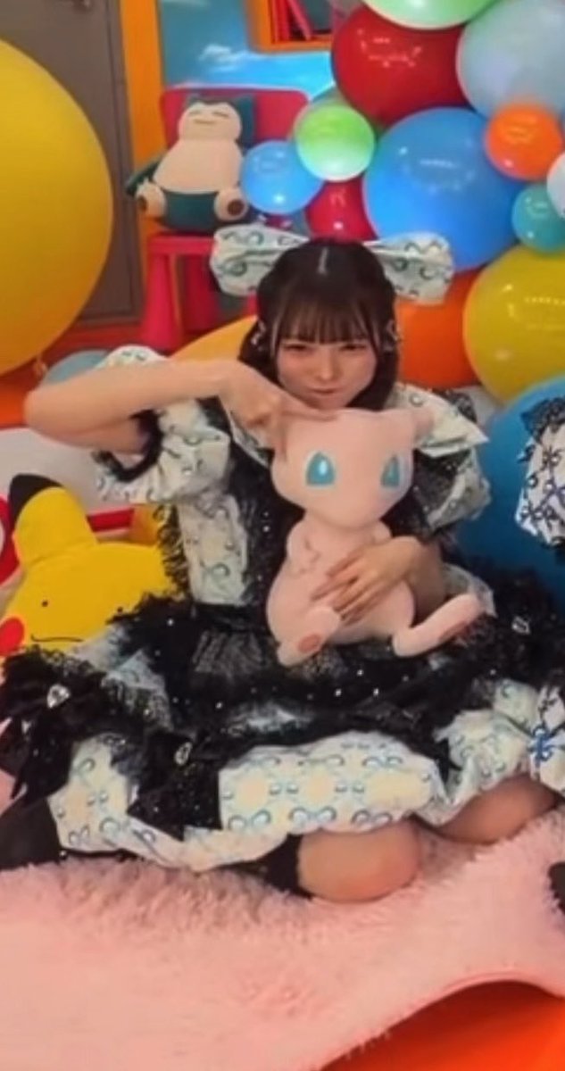A young woman in a black and white frilly dress with star-patterned hair accessories sits surrounded by colorful balloons, holding a pink Mew plush toy while making a cute pose.