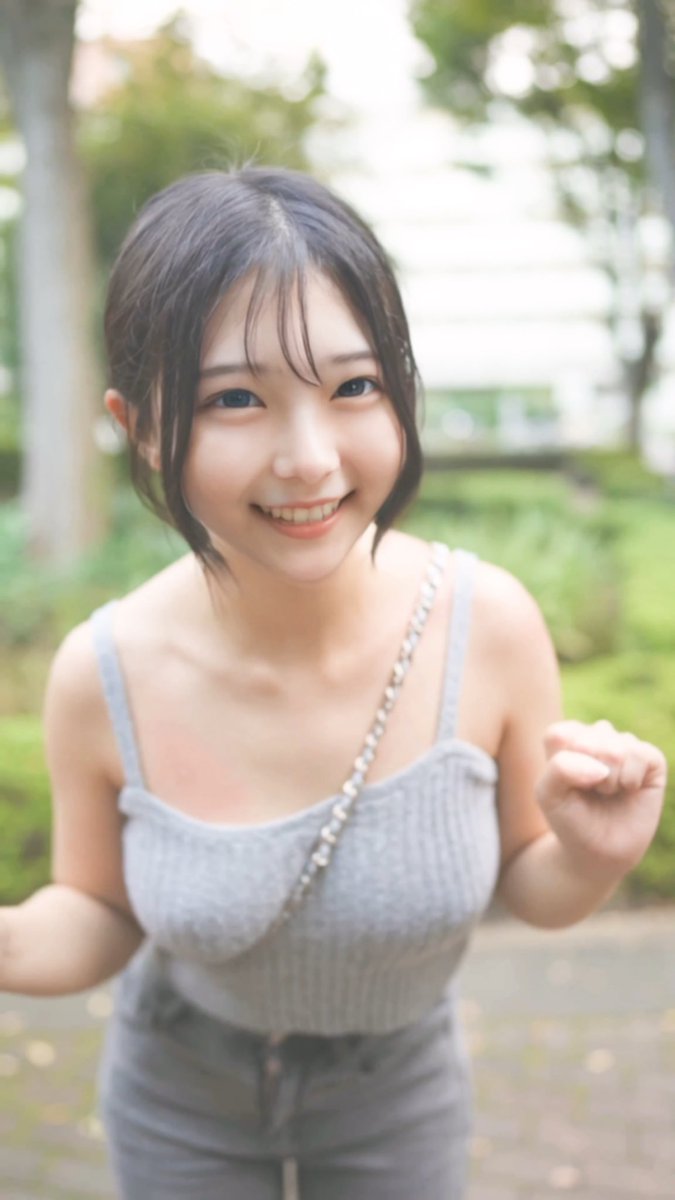 A portrait of a smiling young woman with dark hair in a bob cut, wearing a light blue ribbed tank top and a silver chain strap, standing outdoors against a blurred green background.