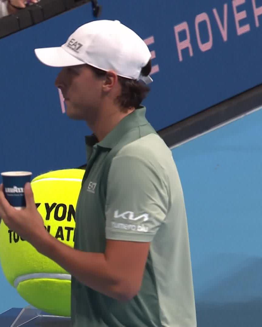 A young male tennis player in an "EAT" cap and light green polo shirt (with "KIA" and "TURKPRO" logos) holds a paper cup and stands near a "YON" oversized tennis ball prop, fitting the "#japanopentennis" setting and referencing the Darderi player's signature look or habit.