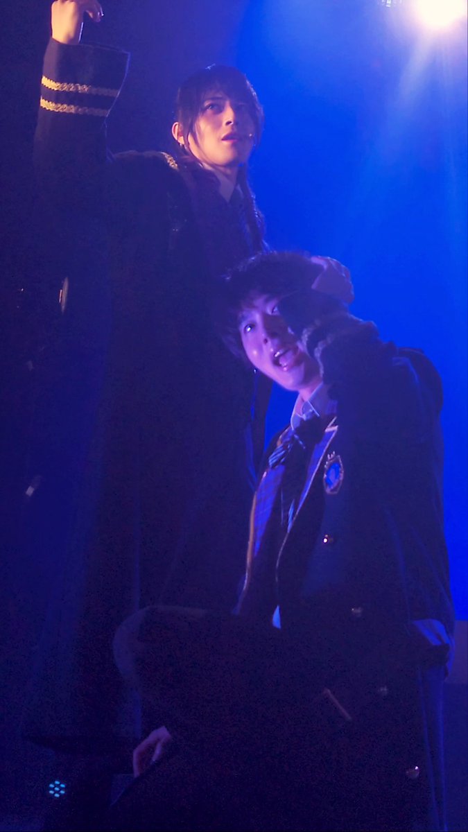 Two performers on a dimly lit stage bathed in blue lighting, one standing tall in the background while the other crouches in the foreground wearing a dark uniform jacket.