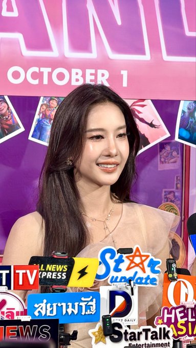 A woman with long dark hair smiles at a media event, with a pink backdrop reading "OCTOBER 1" and multiple news microphones including "TV Channel News," "Star Update," and "StarTalk Entertainment" in the foreground.