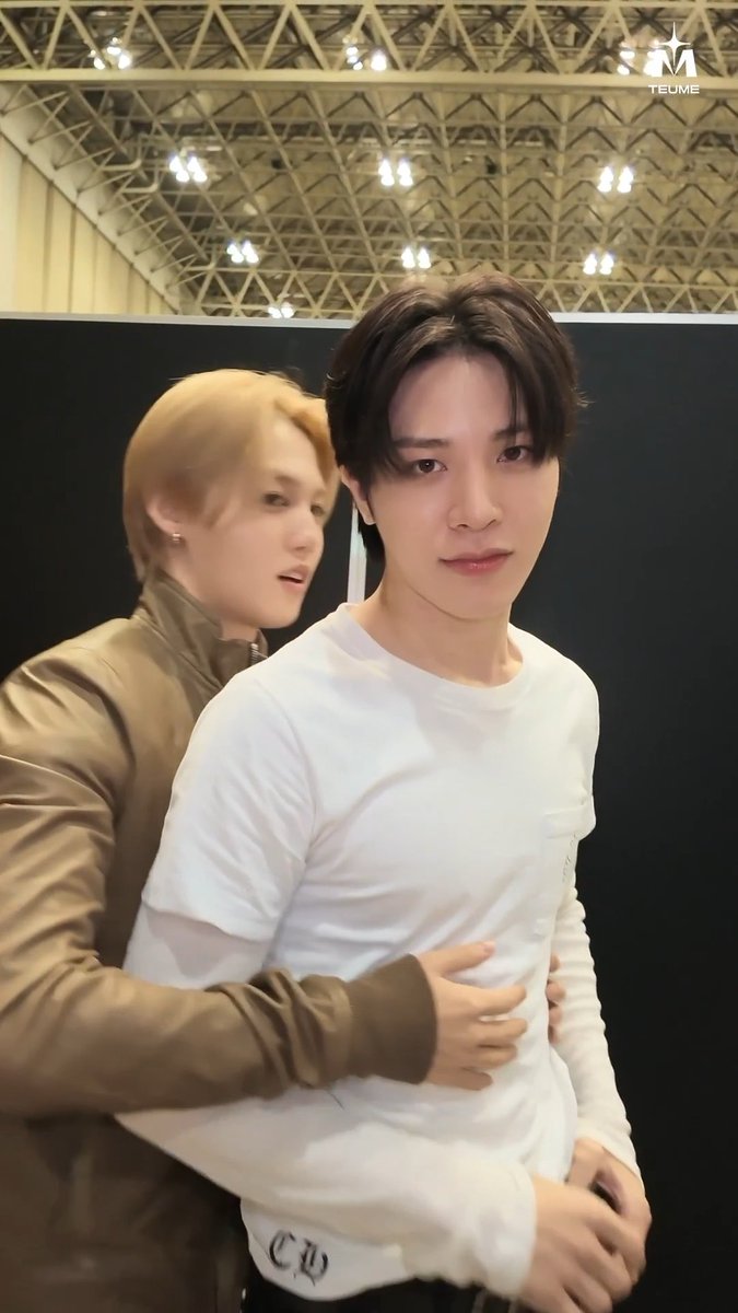 Two young men pose playfully indoors, with one in a brown leather jacket hugging from behind the other who is wearing a white long-sleeve shirt, with a 'TEUME' watermark in the corner.