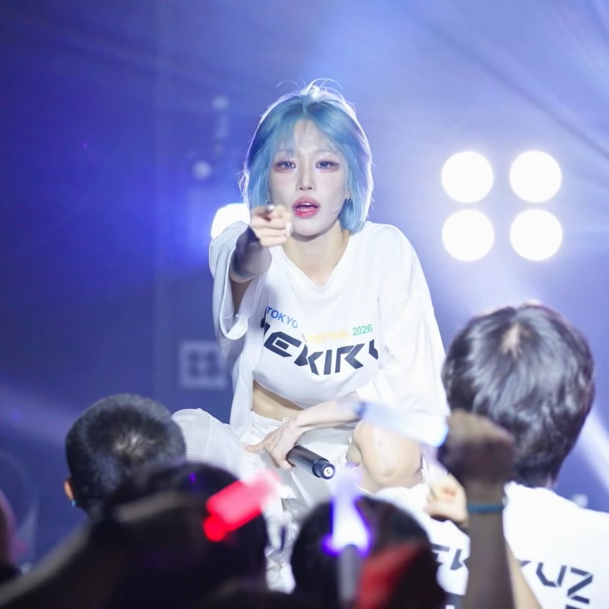 A performer with blue hair wearing a white 'TOKYO IDOL FESTIVAL 2026' t-shirt points dramatically at the camera while on stage.