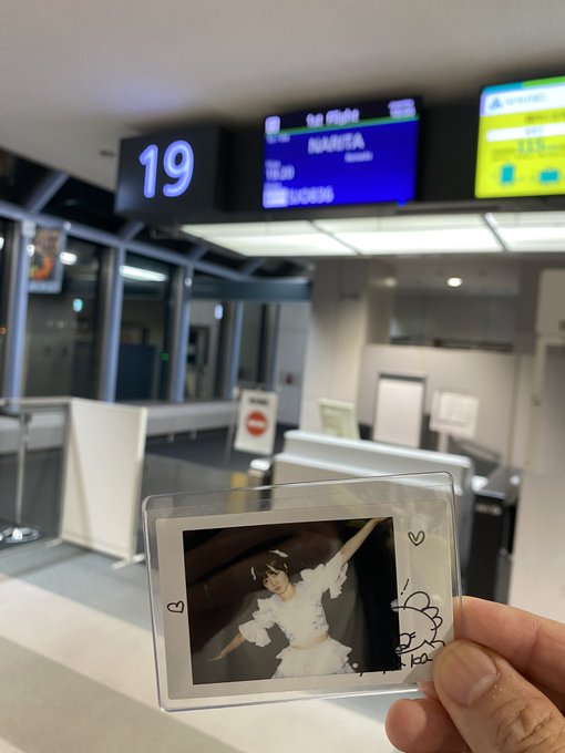 A hand holds up a clear photocard sleeve containing a photo of a woman, taken at an airport gate area displaying "19" and flight information screens in the background.