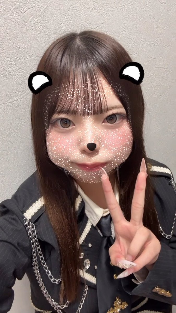 A selfie of a young woman with long brown hair wearing a dark pinstriped jacket with chain details, posing with a peace sign while a digital bear filter with ears, nose, and sparkle overlay is applied to her face.