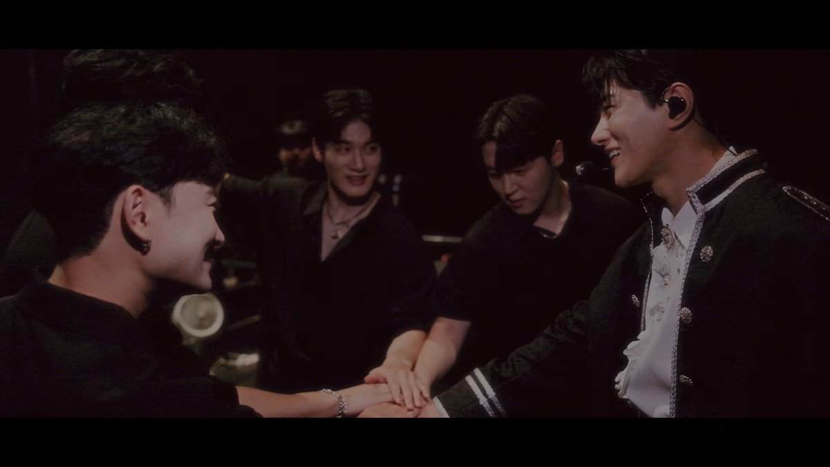 Four young men in dark formal wear are shown backstage or on stage, smiling and placing their hands together in a team huddle.