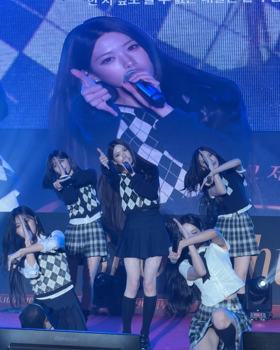 Five female K-pop performers on stage in matching schoolgirl-style outfits with argyle vests and plaid skirts, performing in front of a large screen displaying a close-up of a singer holding a microphone.