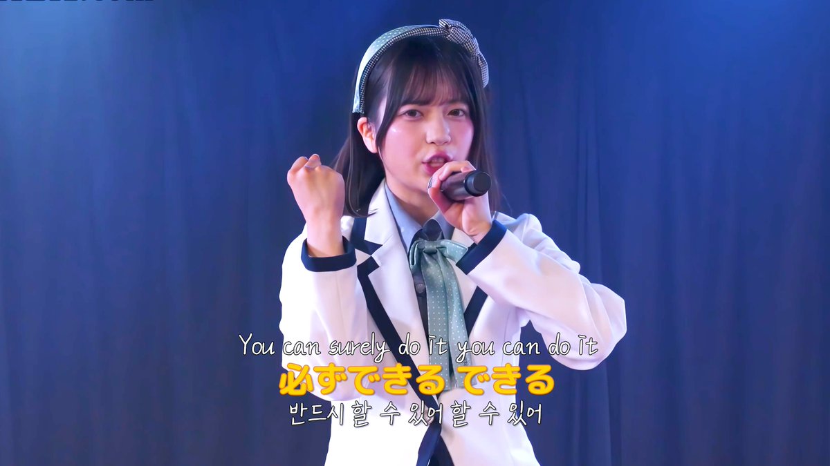 A young woman in a white blazer with a green polka-dot bow and headband singing into a microphone, with subtitles in English, Japanese ('必ずできる できる'), and Korean.
