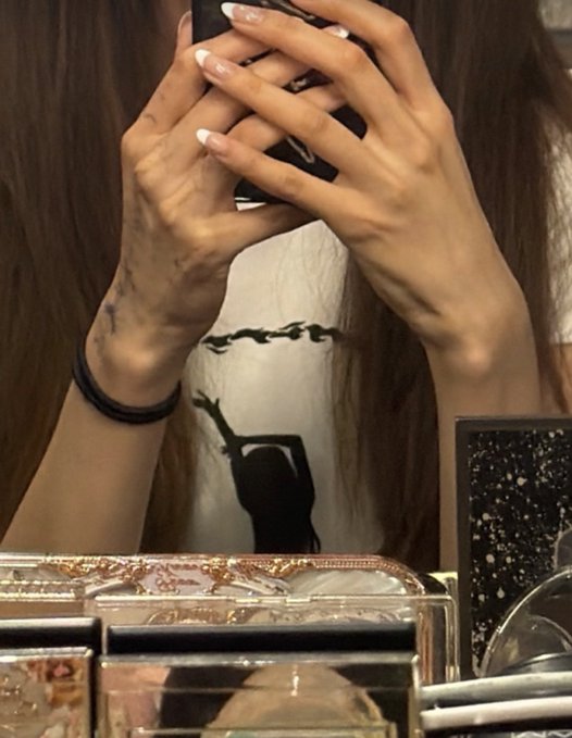 A mirror selfie showing a person's hands holding a phone, with French-manicured nails, a small wrist tattoo, black hair bands, and a Marilyn Monroe graphic tee visible.