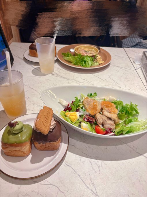 A meal on a marble table featuring a large salad with meat, two drinks, and a plate of decorated pastries including one with kiwi and chocolate toppings.