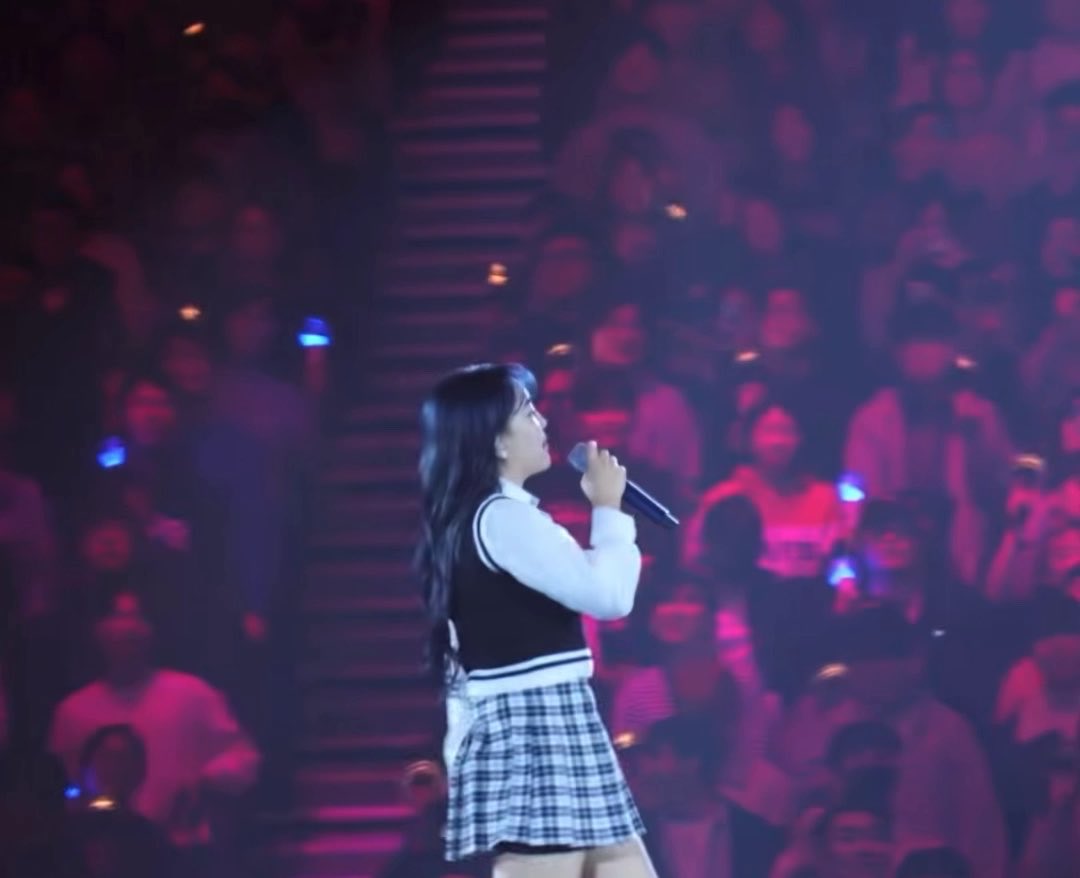 A female performer with long dark hair sings into a microphone on stage wearing a school uniform-style outfit, with an audience visible in the dimly lit background.