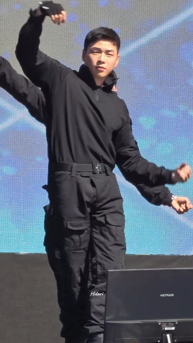 A young man dressed in an all-black outfit including a jacket and belt performs on stage against a blue backdrop, with a monitor displaying 'VICTRACK' in the foreground.