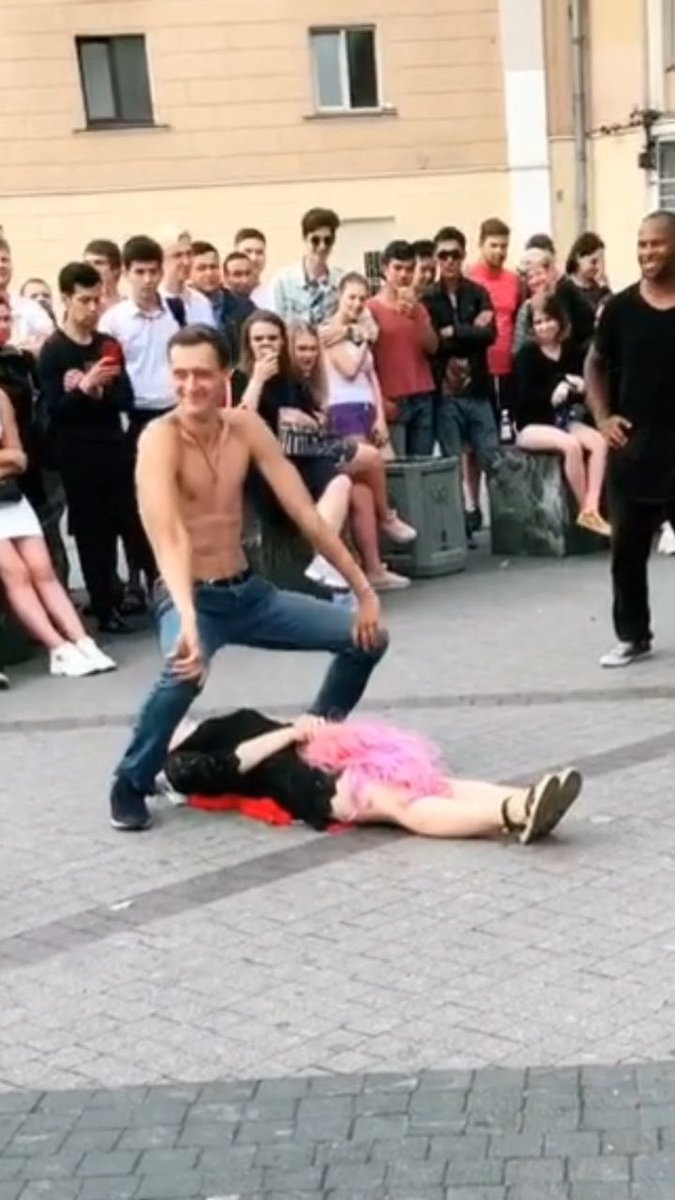 A shirtless young man in jeans squats smiling over a person in a black top and pink tutu lying on the ground, while a crowd of onlookers watches on a city street.
