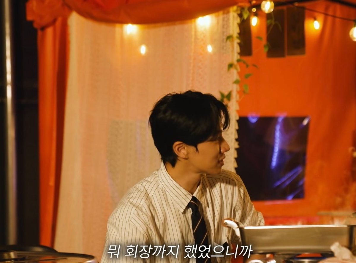 A young Asian man in a striped button-up shirt and tie, shown in profile against a warm-lit background with orange curtains and string lights, with Korean subtitles reading '뭐 회장까지 했었으니까' (Since I was even the chairman).
