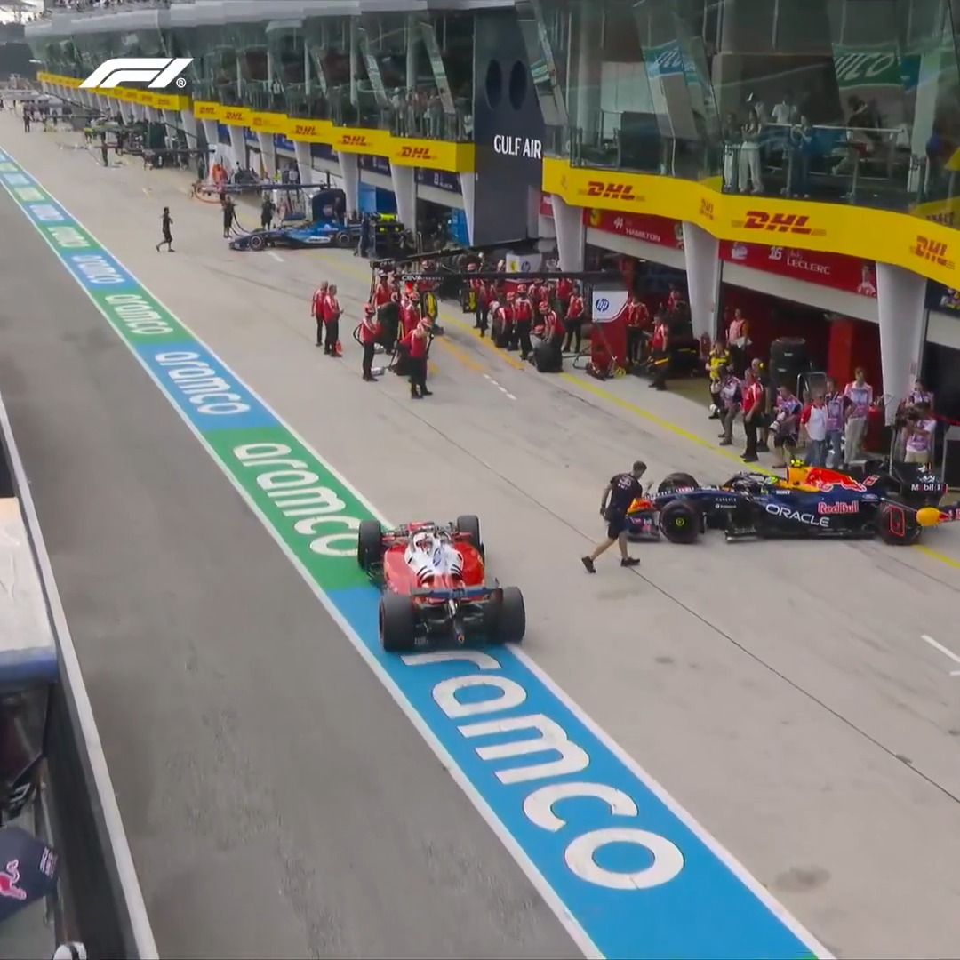 A red Alfa Romeo (likely driven by Zhou Guanyu) navigates the pit lane as a Red Bull car on the right passes by, with a person walking between the two vehicles—aligning with the caption of a close encounter during pit lane action at the Bahrain Grand Prix. Visible signage includes "GULF AIR," "DHL," "aramco," "ORACLE Red Bull," "hp," and "LECLERC," along with the F1 logo in the top left.