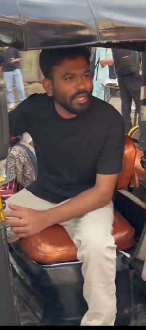A bearded man in a black t-shirt and white pants sits on the back seat of an auto-rickshaw or tuk-tuk, speaking while seated on the orange cushion.