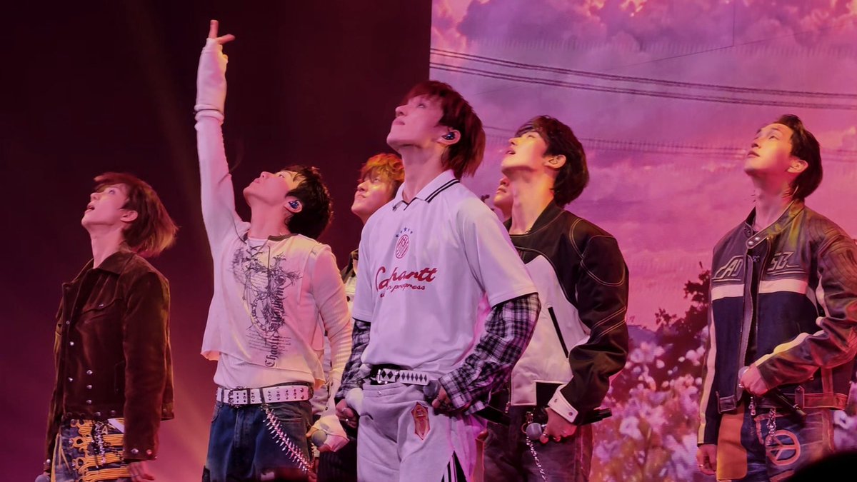 A K-pop boy group performs on stage under pink lighting, looking upward with one member pointing toward the sky.