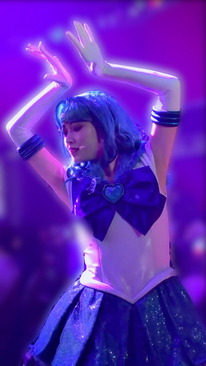 A performer dressed as Sailor Mercury, in a blue and white sailor uniform with a heart brooch, posing with arms raised against a purple background.