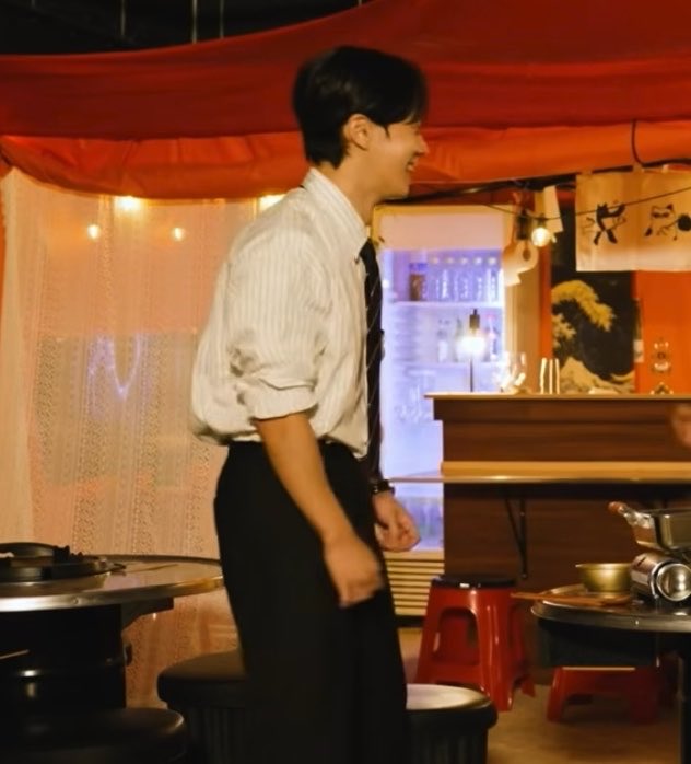 A young man wearing a white striped shirt, tie, and black pants smiles while standing in what looks like a restaurant or bar setting with red fabric overhead and a counter in the background.